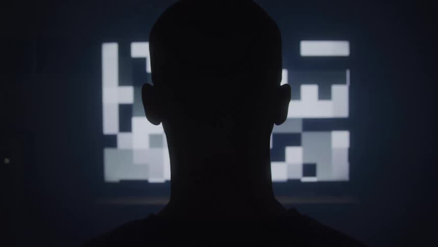 A blad man stands in darkness staring intently at a glowing screen filled with abstract pixelated blocks