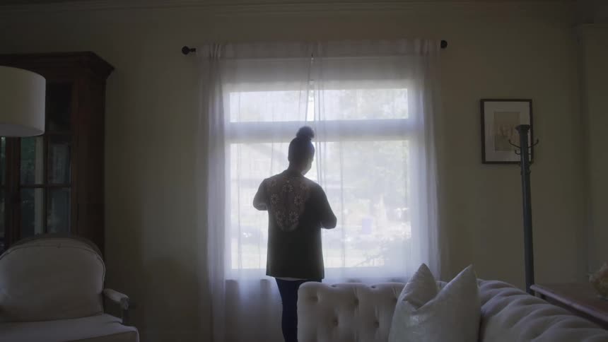 An older adult woman with her hair in a bun wearing a patterned top and dark pants stands with her back to the camera looking out a large window with sheer curtains in a living room furnished with a couch and armchair