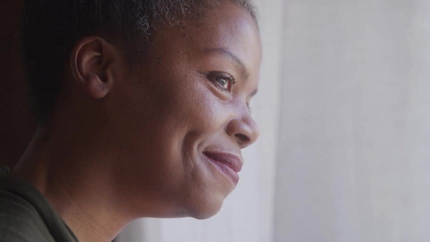 An older adult woman with short grey hair smiles and looks thoughtfully off to the right side of the frame with her face partially illuminated by soft light