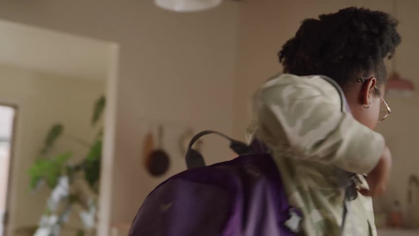 A black female teenager carries her purple bag over her shoulder as she stands and walks away from the living room during daytime