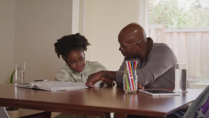 An adult man likely an instructor wearing glasses and a grey shirt sits at a wooden table helping a young female student with curly hair and glasses as she writes in an open book with a glass of water and colorful pencils on the table