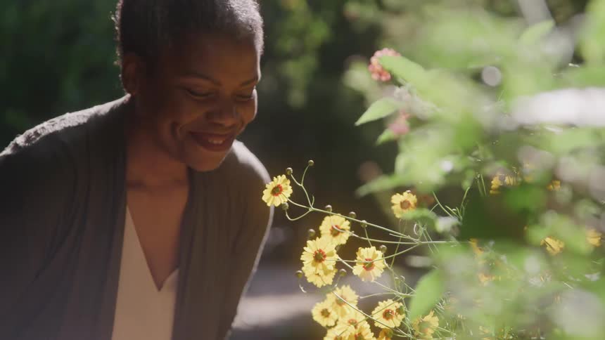 An older adult woman with short grey hair wearing a green cardigan and a light colored top smiles and looks down at a cluster of bright yellow flowers in a sunny outdoor setting
