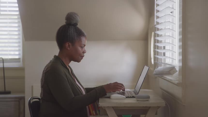 An older adult woman with her hair in a bun wearing a green cardigan sits at a desk and types on a laptop with blinds on the window and a smart speaker nearby