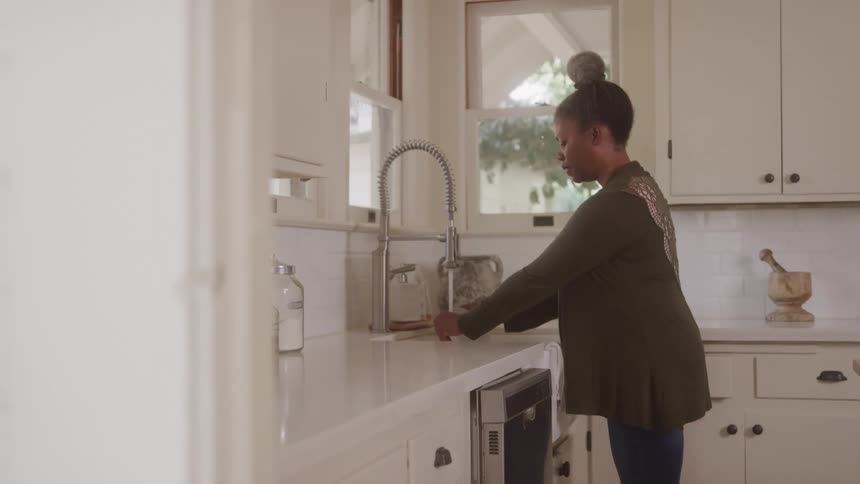 A woman stands at a kitchen counter near the sink washing a dish in a bright cozy kitchen