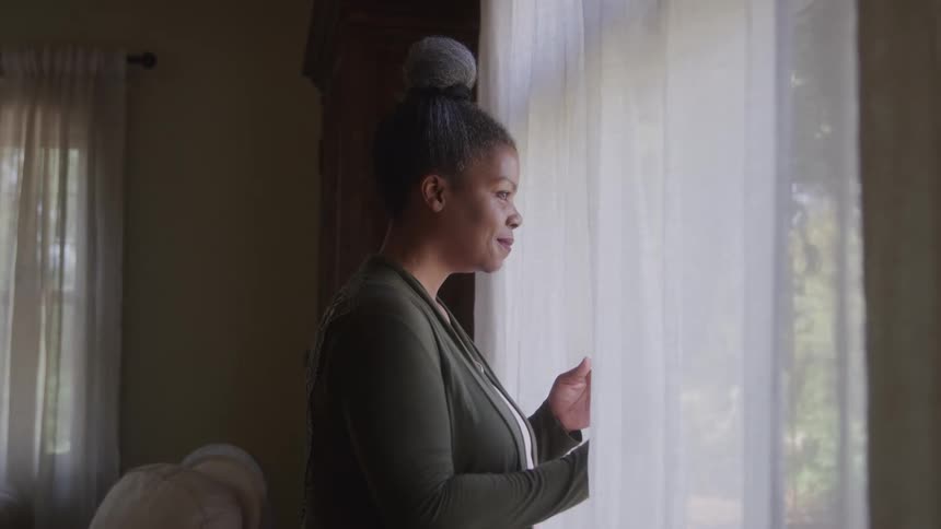 A woman walks toward a window with white curtains at midday looking outside through the curtain with a smile on her face