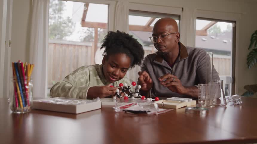 A girl and an adult man are sitting at a table with science supplies the man helps the girl put together a molecular model from a science kit