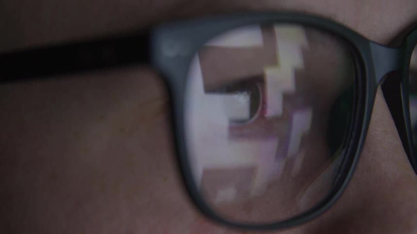 A close view of an adult persons face shows their eye through glasses with square pixelated reflections on the lenses