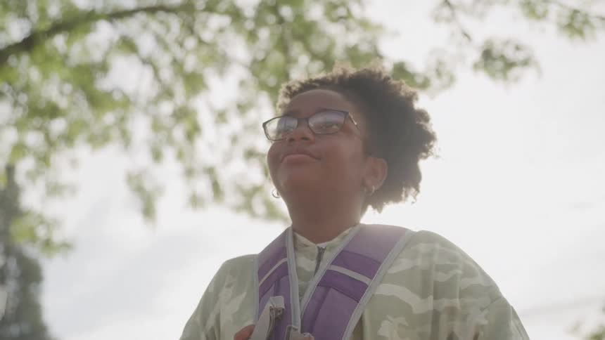 A young adult woman with curly hair wearing glasses and a camouflage shirt with a purple strap over her shoulder looks upwards with a pleasant expression as bright sunlight shines through the green leaves of trees