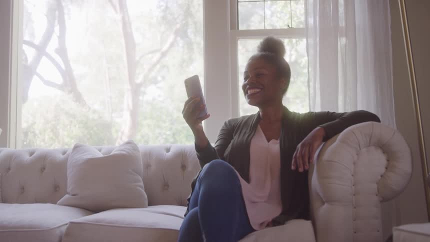 A smiling woman is seated on a white sofa holding a phone and waving at the screen likely during a video call the scene is bright and suggests a casual happy moment