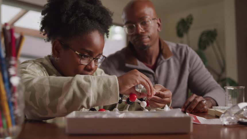 Father sitting next to his daughter watching her build a chemical structure with colored objects on a table