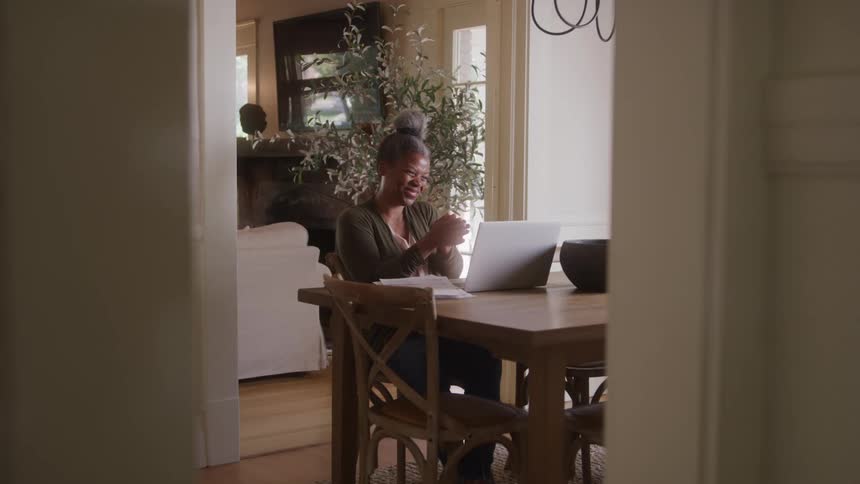 Woman sitting at a table waves at the laptop in front of her while she talks during a video call