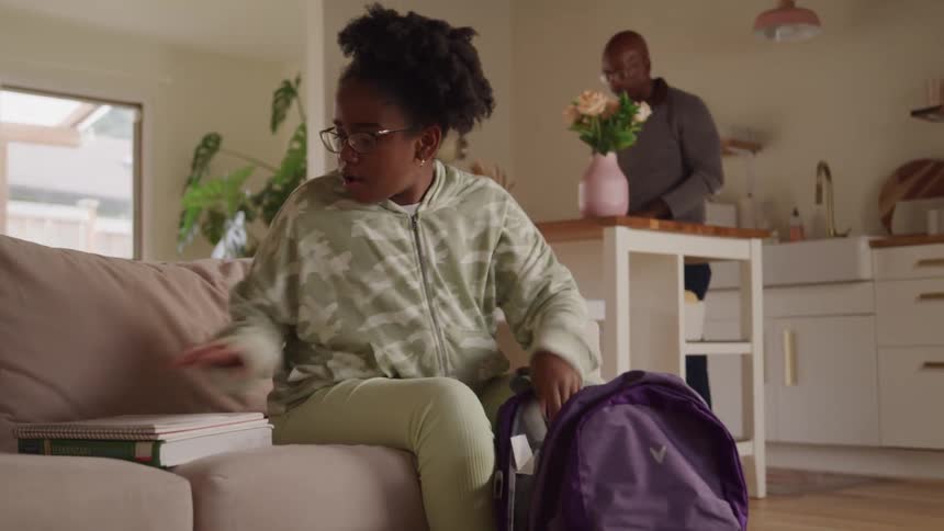 A female black teenager puts her books and writing materials into her backpack while sitting on the couch in the living room during daytime