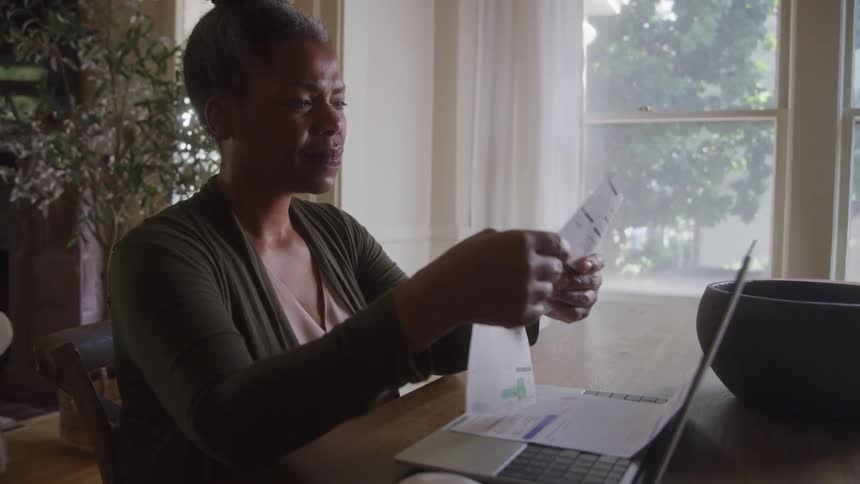 An older adult woman with her hair in a bun wearing a green cardigan sits at a table looking at papers with a laptop a computer mouse and a dark bowl in front of her and a large window looking out at trees in the background