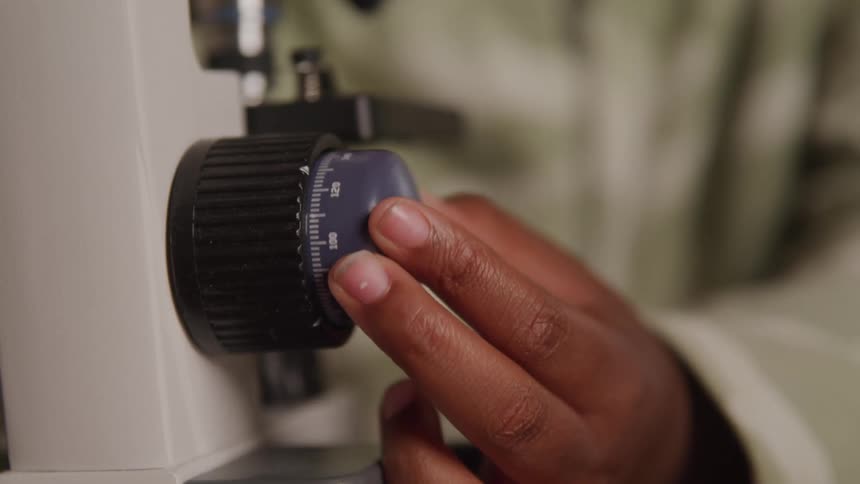 A close view shows a persons hand adjusting a black dial on a piece of scientific equipment likely a microscope