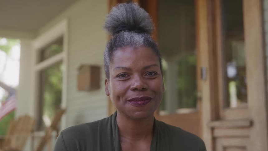 A smiling woman with grey hair styled in a high bun looks directly at the camera standing outdoors with a blurred background of a house porch she is wearing a dark green top