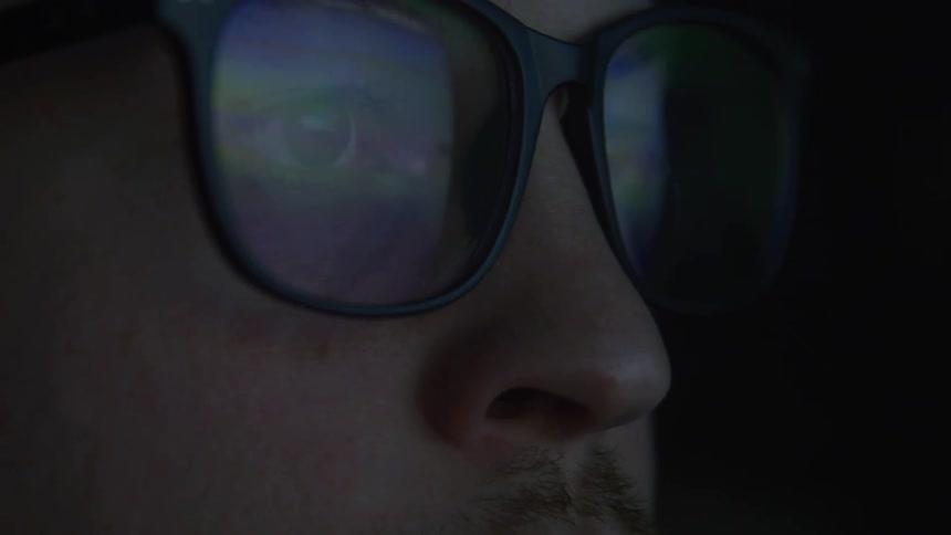 A closeup reveals a person wearing glasses with reflections of digital screens flickering across the lenses the background is dark emphasizing the glow on the glasses suggesting the person is deeply immersed in viewing content on a device