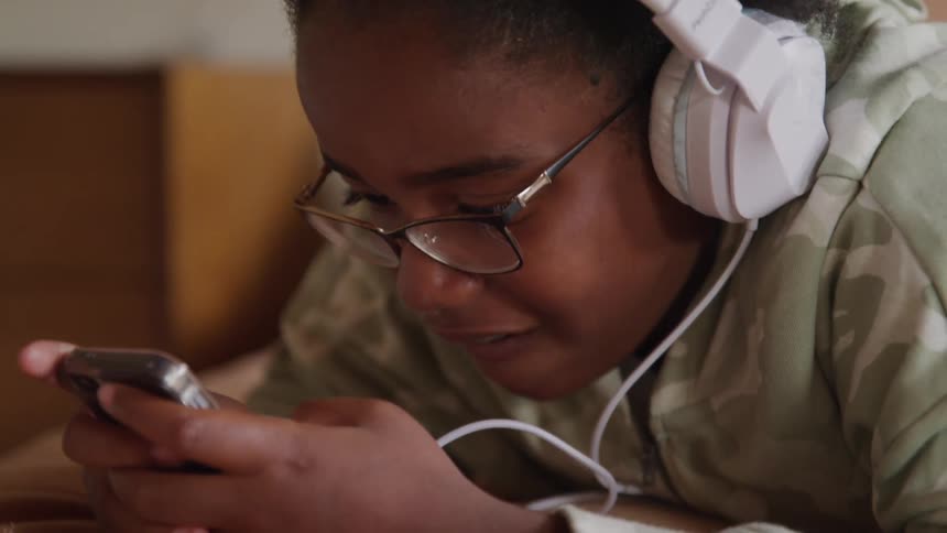 Close up of a black female teenager using her phone while lying down indoors during daytime