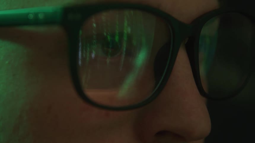 A close view of an adult persons face seen in profile shows their eye through glasses with green digital code reflecting on the lenses