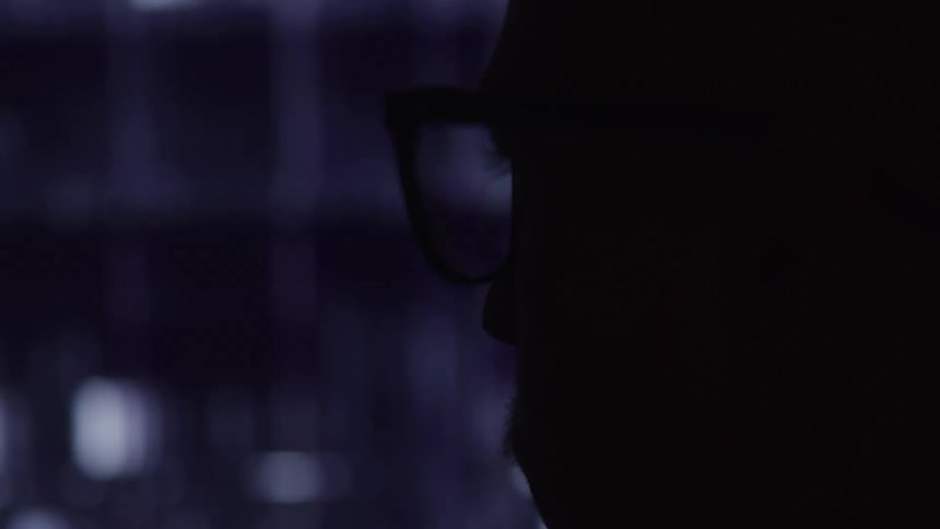 A closeup shot shows a persons head in dark silhouette with visible glasses looking towards a blurred background of horizontal light and dark lines possibly reflections on a screen or window the lighting creates a stark contrast highlighting the ou