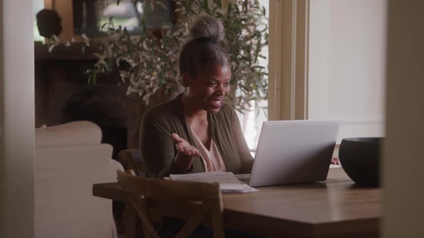 A woman with her hair in a bun is sitting at a wooden table in front of a laptop she smiles and gestures with her hands as she appears to be on a video call