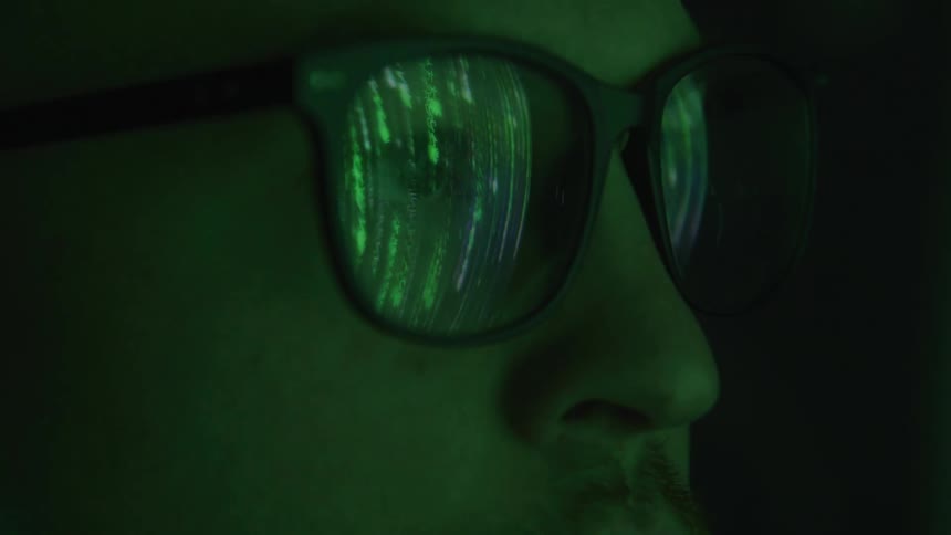 A close view of a persons face seen in profile shows green digital code reflecting on the lenses of dark framed glasses