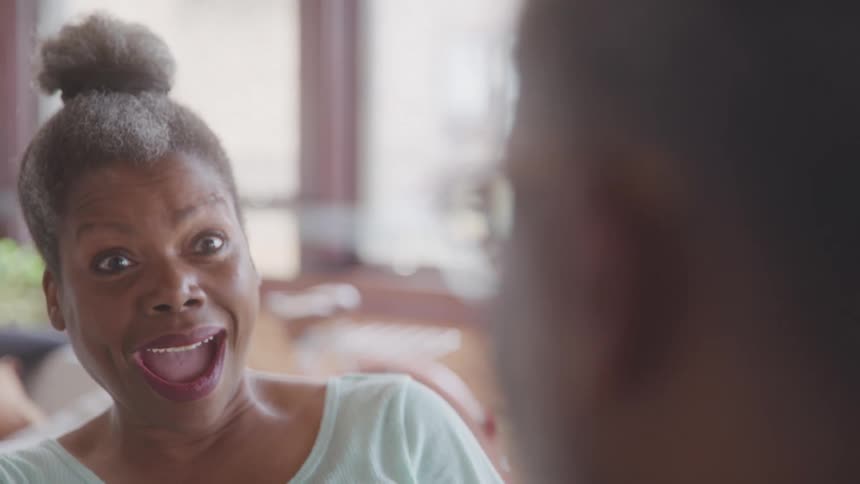 An older adult woman with her grey hair in a bun wearing a light colored top holds up a red playing card with an excited open mouthed expression then smiles as she looks at another person whose shoulder is blurred in the foreground
