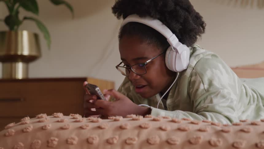 A kid lies on the bed wearing a headset and glasses scrolling through her phone