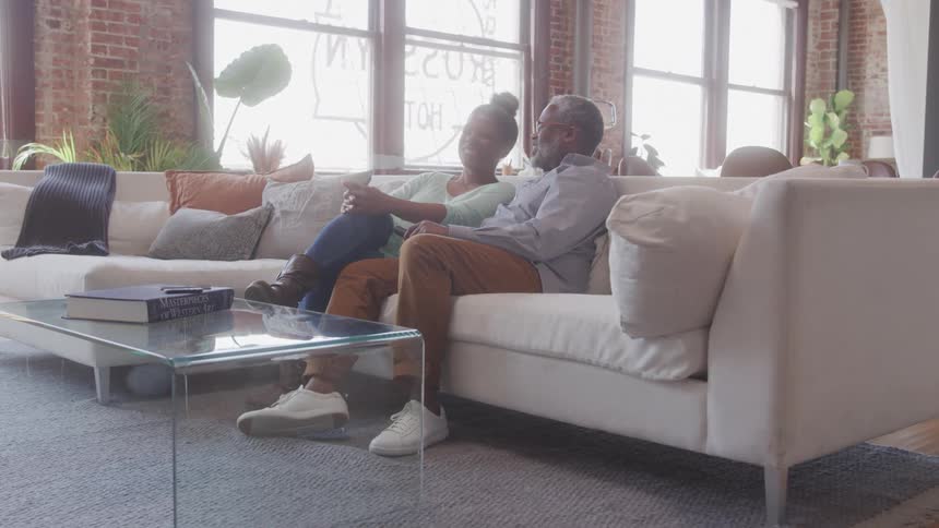 An older adult couple sits comfortably on a light colored sofa in a living room smiling and looking out a large window with exposed brick walls a coffee table and plants completing the home setting