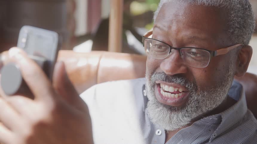 An elderly man with gray hair and a gray beard sits outdoors at midday wearing glasses and enjoying a video call on his phone