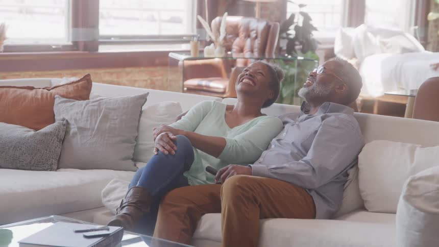 A man and woman sit on a couch in the living room at midday laughing and enjoying their time together