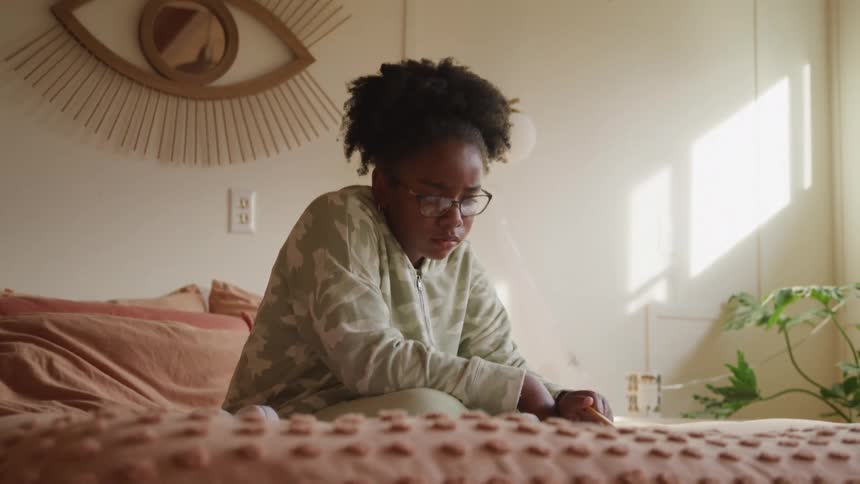 A child sits one the bed and writes with a pencil