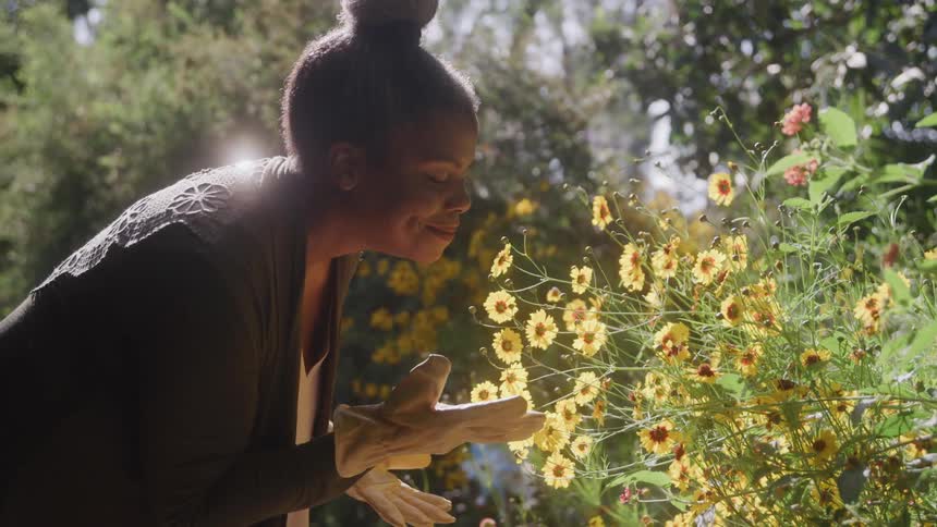A black woman in gardening gloves gently inhales the scent of vibrant yellow flowers in a sunlit garden