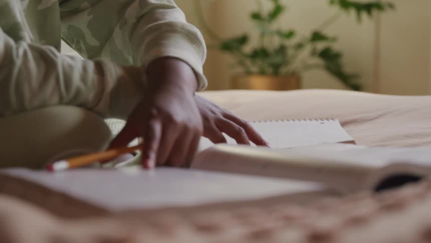 A person wearing lightcolored clothing is seated on a bed holding a pencil and writing in a notebook or book another open book lies next to them with a green potted plant visible in the blurred background