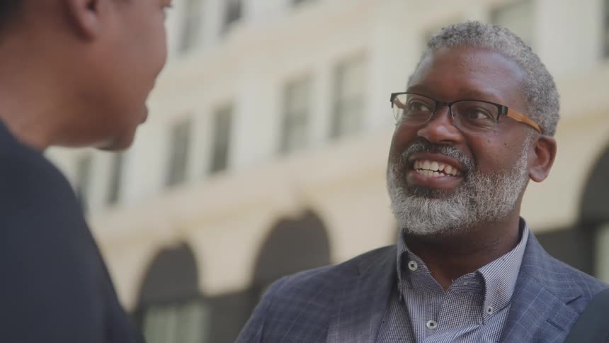 An older adult man with grey hair and a beard wearing glasses and a suit jacket smiles widely while looking at another person whose back is to the camera as they converse outdoors in front of a building