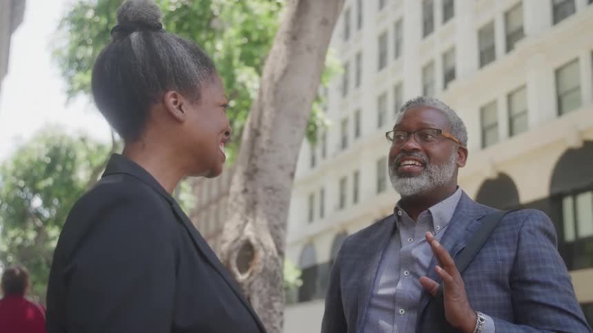 An older man and an older woman both in business attire converse on a city street with the man gesturing as they talk in front of a building and trees