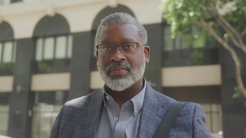 A mature man with a grey beard and glasses is standing outdoors looking directly at the camera with a serious but pleasant expression he is wearing a suit jacket and a blue striped shirt with a building visible in the blurred background