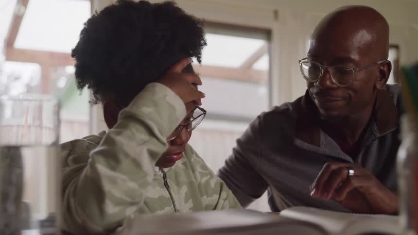 A teenage girl sits at the dining table reading and looking frustrated while her dad sits beside her encouraging her with a tap on the back as she rests one hand on her face