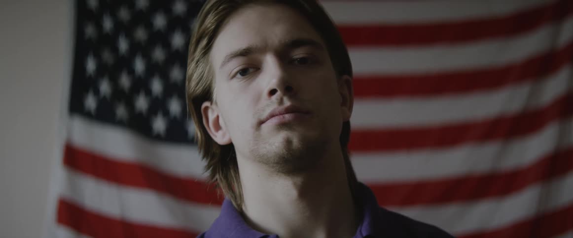 A man standing and posing in front of a wall with an american flag in the background