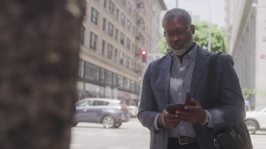 An older adult man with grey hair and a beard wearing glasses and a business suit with a shoulder bag looks at his smartphone while standing on a city sidewalk with buildings and parked cars