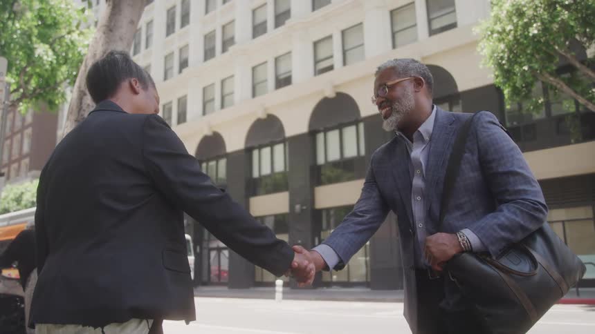 A man wearing a suit and holding a handbag shakes hands with a woman in the middle of the street at midday both smiling at each other