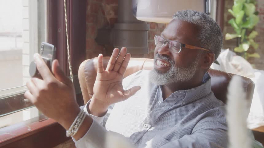 An older adult man with grey hair and a beard wearing glasses and a light colored collared shirt sits in a brown leather chair near a window smiling and waving at a smartphone held in his hand as if on a video call