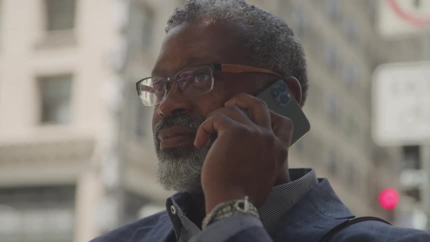 A middleaged man with a gray beard and glasses is seen on a city street holding a smartphone to his ear he appears to be engaged in a phone conversation with urban buildings visible in the blurred background