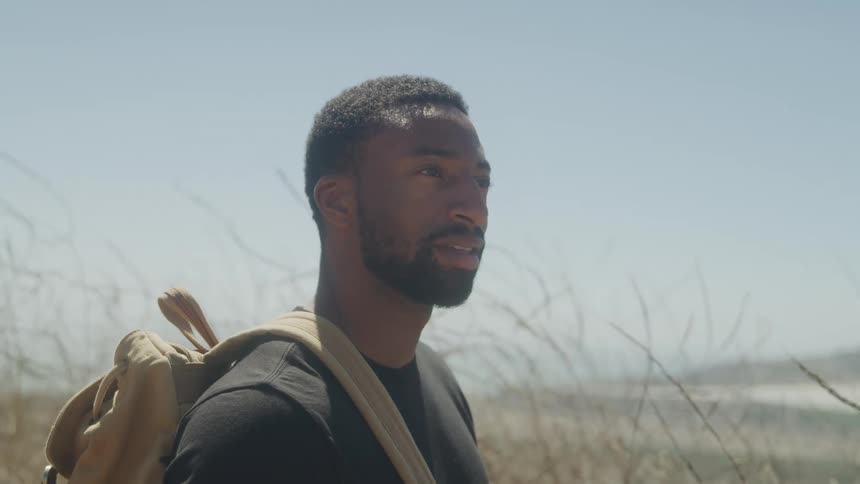The face of a young man wearing a black shirt and a beige backpack in an outdoor setting with vegetation behind him