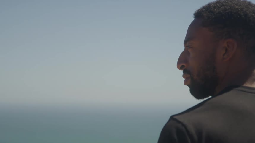 A young adult man with short hair and a beard wearing a dark t shirt looks out towards a wide body of water and the horizon