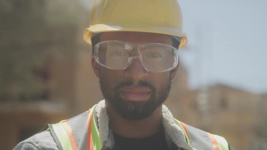 A young adult man likely a construction worker or supervisor wearing a yellow hard hat safety glasses and a safety vest over a jacket looks directly at the camera with a subtle smile