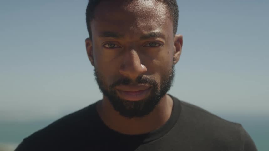 A young adult man with a beard and short hair wearing a dark shirt looks down then directly at the camera with a serious expression with a blurred outdoor background