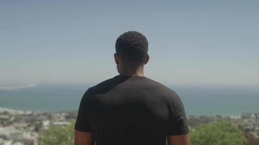 A young adult man with short hair wearing a dark t shirt stands with his back to the camera looking out at a wide body of water with a distant city and bright sky