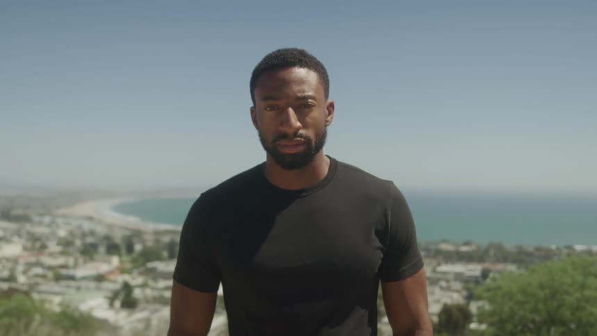 A man with a beard and short hair wearing a dark t shirt looks directly at the camera with a neutral expression standing outdoors with a vast coastline and urban area in the background