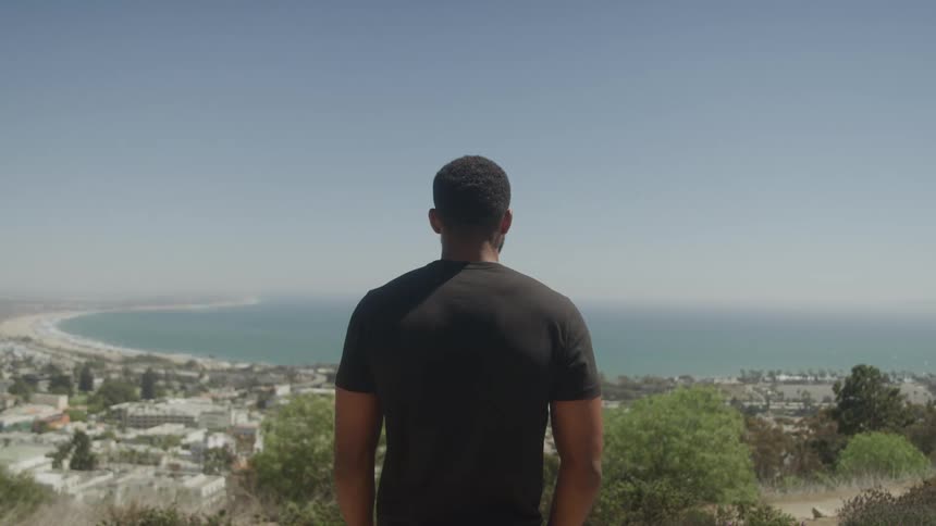 A person is seen from behind standing on a hill overlooking a city and a vast body of water with a clear sky above the scene captures a panoramic view of the coastline and urban landscape