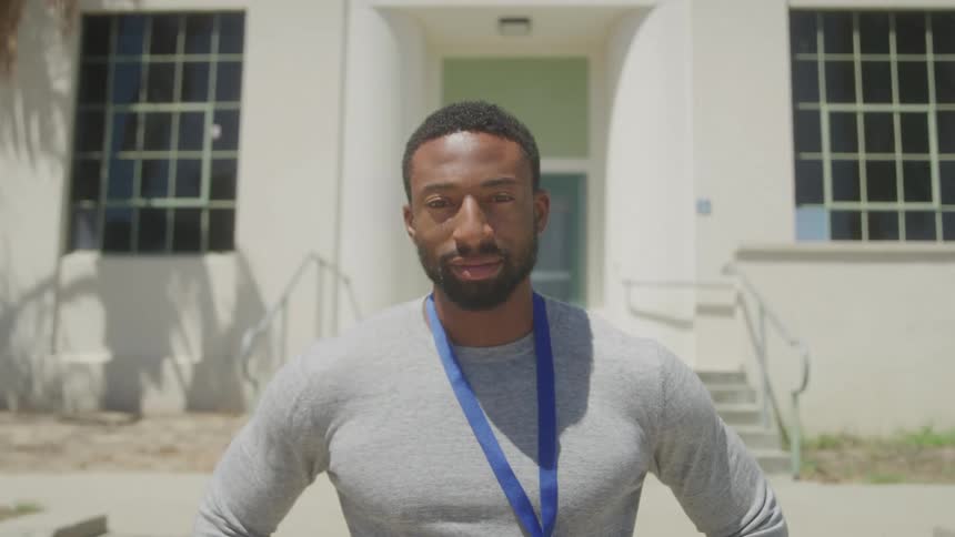 A man with a beard and short hair wearing a grey shirt and a blue lanyard looks to his right then turns to face the camera and smiles standing outdoors in front of a white building with large windows